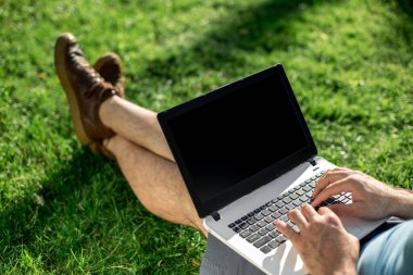 Cropped shot of man using laptop with blank screen while sitting on green grass. Freelance works in the open air in the park