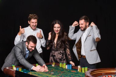 Rulet masasının arkasında bir grup genç siyah arka planda. Genç adam zafere seviniyor.