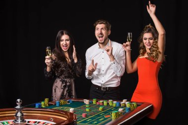 Siyah arka planda rulet masasının arkasında iki genç kadın ve erkek. Duygusal oyuncular