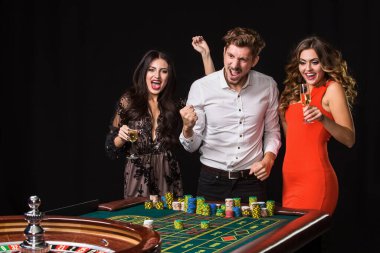 Siyah arka planda rulet masasının arkasında iki genç kadın ve erkek. Duygusal oyuncular