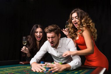 Siyah arka planda rulet masasının arkasında iki genç kadın ve erkek. Duygusal oyuncular