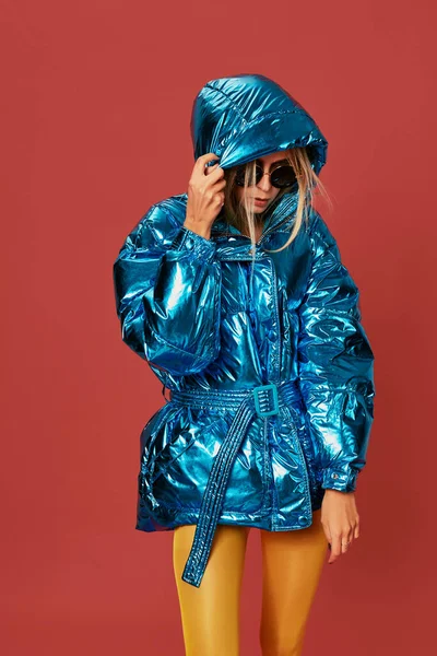 Close up fashion photo of young woman covering herself with hood. She wears blue oversized down jacket in studio on red background
