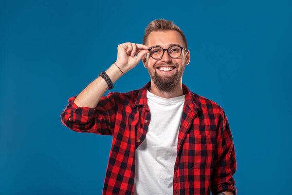 Emotional and people concept: young bearded man in checkered shirt. Hipster style. Studio shot on blue background. Copy space. The guy adjusts his glasses