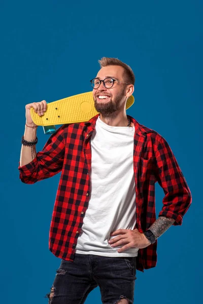 カリスマ的な陽気な若いひげを生やした男は 彼の手の中に黄色のスケートボードで チェッカーシャツ 白いTシャツと眼鏡を着ていた スタジオは青い背景で撮影しました ヒップスター — ストック写真