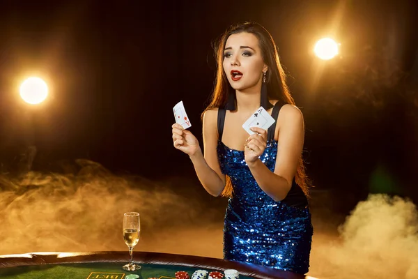 青い光沢のあるドレスの若い美しい女性は 高級カジノのポーカーテーブルの近くにポーズをとります 女選手 カード チップ アルコール ダイス ギャンブル カジノ それは女性のエンターテイメントとしてです — ストック写真