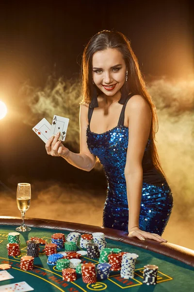 青い光沢のあるドレスの若い美しい女性は 高級カジノのポーカーテーブルの近くにポーズをとります 女選手 カード チップ アルコール ダイス ギャンブル カジノ それは女性のエンターテイメントとしてです — ストック写真