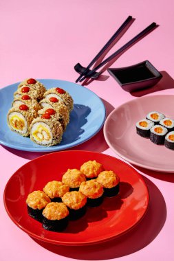 Tempura karidesli lezzetli suşi, kremalı maki suşi ve pembe arka planda soya soslu renkli tabaklarda servis edilen somon balığı. Popüler Japon mutfağı