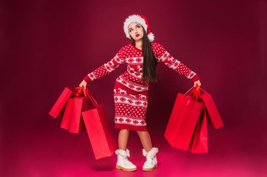 Çekici kadın iki eliyle bir sürü hediye paketi tutuyor. O ayakta duruyor ve gülüyor. Noel Baba şapkalı genç bayan mutlu bir şekilde bekliyor. Kırmızı arkaplanda izole