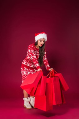 Çekici kadın iki eliyle bir sürü hediye paketi tutuyor. O ayakta duruyor ve gülüyor. Noel Baba şapkalı genç bayan mutlu bir şekilde bekliyor. Kırmızı arkaplanda izole