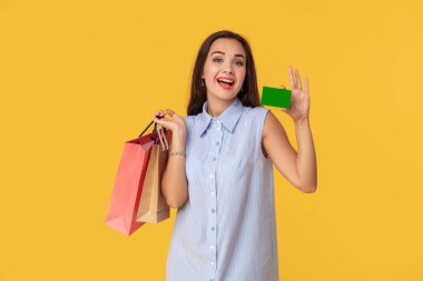 Farklı alışveriş paketleri ve kart üzerinde sarı arka plan izole elinde tutarken gülümseyen uzun kahverengi saçlı elbiseli trendy kadın 20'li yaşlarda. Alışveriş. Satılık