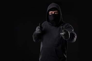 Siyah arka planda kameraya doğrultulmuş silahlı maskeli bir soyguncu. Silahlı bir adam.