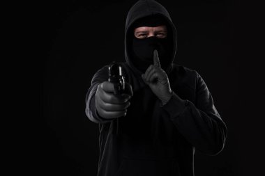 Siyah arka planda kameraya doğrultulmuş silahlı maskeli bir soyguncu. Silahlı bir adam.