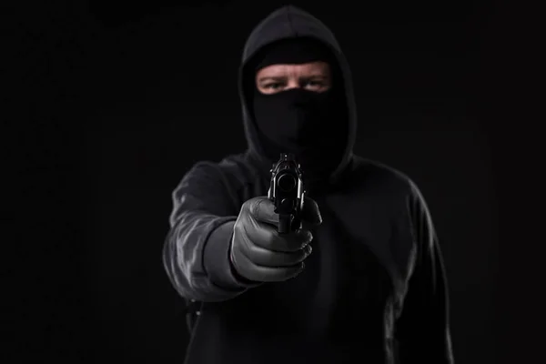 Masked robber with gun aiming into the camera against a black background. Man with a gun