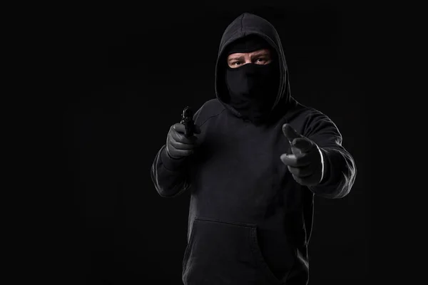 Masked robber with gun aiming into the camera against a black background. Man with a gun