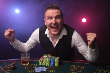 Beyaz gömlekli ve siyah yelekli şık, olgun bir adam kumarhanede kırmızı ve mavi renkli bir adama karşı poker masasında üzerinde cips ve bir bardak viski ile poz verdiği için çok mutlu görünüyor.