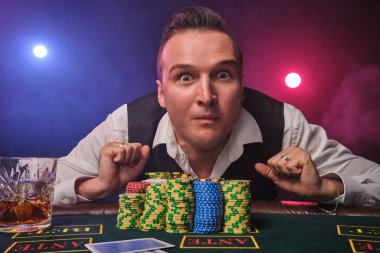 Beyaz gömlekli ve siyah yelekli şık olgun bir adam, kumarhanede üzerinde cips ve bir bardak viski olan bir poker masasında kırmızı ve mavi arka farlara karşı poz verirken şaşırmış görünüyor.