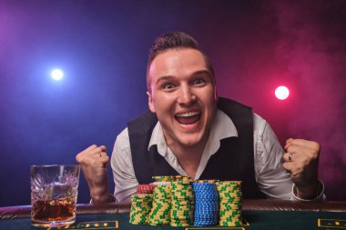 Beyaz gömlekli ve siyah yelekli zarif olgun bir insan kumarhanede üzerinde cips ve bir bardak viskiyle poker masasında kırmızı ve mavi arka farlara karşı poz verirken heyecanlı görünüyor.