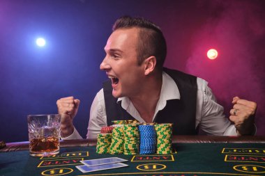 Beyaz gömlekli ve siyah yelekli zarif olgun bir adam kumarhanede üzerinde cips ve bir bardak viskiyle poker masasında poz verirken mutlu görünüyor.