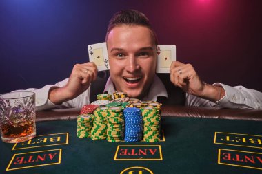 Beyaz gömlekli ve siyah yelekli zarif olgun bir adam elinde iki iskambil kâğıdı tutuyor ve kameraya bakıyor. Poker masasında poz veriyor. Üstünde cips ve bir bardak viski var.