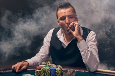 Beyaz gömlekli ve siyah yelekli ciddi bir olgun adam masada oturmuş puro içiyor. Kumarhanede poker oynuyor. Arka planda siyah dumanlı bir ışık altında.