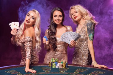 Charming females with a perfect hairstyles and bright make-up, dressed in a golden shiny dresses are posing standing at a gambling table. Poker concept on a black smoke background with pink and blue clipart