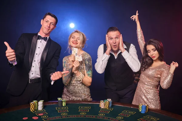 Group of a happy rich friends are playing poker at casino. Youth are making bets waiting for a big win. They are looking overjoyed standing at the table against a red and blue backlights on black