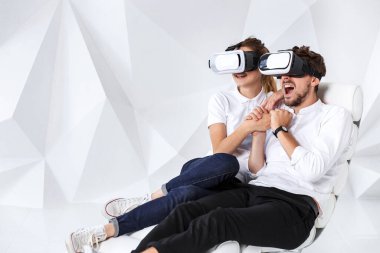 Beyaz duvarlı ve zeminli bir odada beyaz koltukta oturan VR kulaklık takan genç bir çift. Kot pantolon, beyaz spor ayakkabı ve tişört giyen genç bir çift..