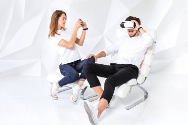 Çift sanal gerçeklikle oynarken eğleniyor. Genç bir adam beyaz duvarlı bir odada rahat bir koltukta oturuyor.