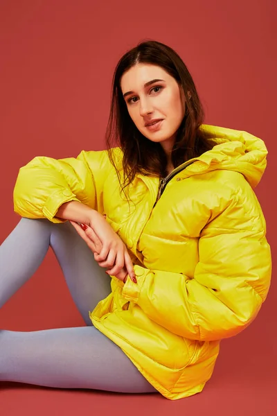 Young dark haired girl sitting on the floor before camera in studio on red background. Fashion image of stylish woman wearing yellow jacket