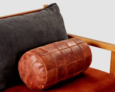 Comfortable cylindrical bolster cushion, hand-stitched from patches of genuine copper-colored leather, for use as stylish decorative accessory on soft sofa