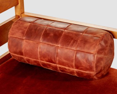 Decorative bolster cushion made of patches of copper-colored leather lying on wooden sofa. Stylish handcrafted furnishing accessory