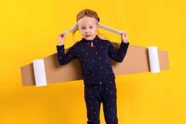 Küçük bir çocuk kız ev yapımı karton uçan kanat, bir zanaat, hayal veya keşif kavram için bir pilot gibi davranan giyiyor. Sarı bir arka plan üzerinde fotoğraf stüdyosu