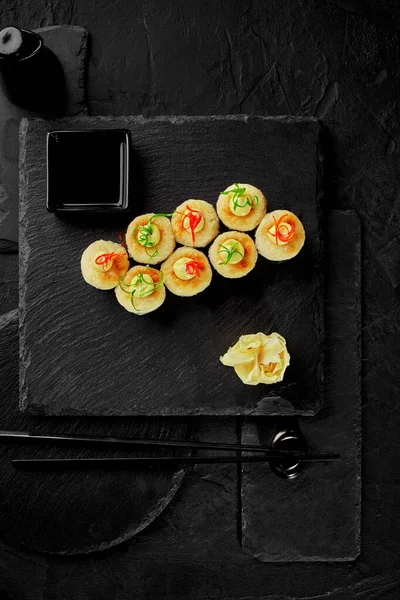 Stock image Top view of baked maki sushi rolls topped with melted cheese, japanese mayonnaise drops, colorful thin shaving of scallions and chili pepper served with gari and soy sauce on black slate board