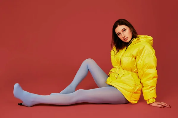 若い褐色の髪の女の子は赤い背景のスタジオでカメラの前に床に座って 黄色のジャケットを着ているおしゃれな女性のファッション画像 — ストック写真