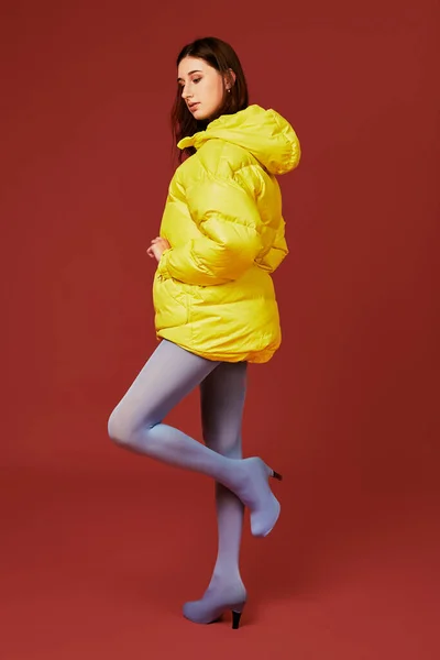 赤を背景にスタジオでカメラの前にポーズをとる若い黒髪の少女のイメージ 黄色のジャケットを着たスタイリッシュな女性のファッションイメージ サイドビュー — ストック写真