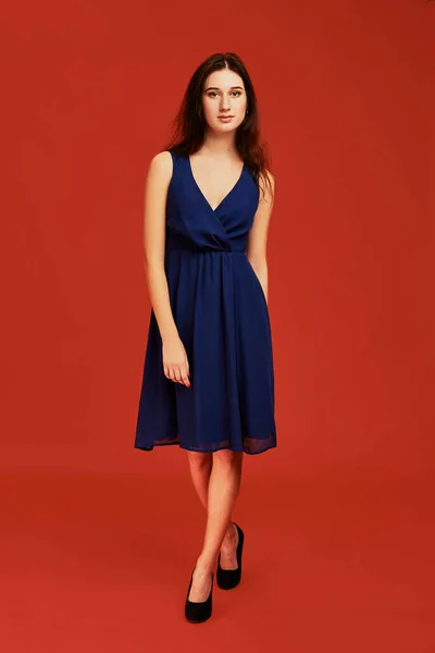 stock image Beautiful young brunette woman in elegant blue cocktail dress and black high heels is posing for the camera. Front view. Full length studio shot on red background