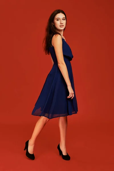 stock image Beautiful young brunette woman in elegant blue cocktail dress and black high heels is posing for the camera. Side view. Full length studio shot on red background