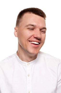 Beyaz gömlekli yakışıklı genç adam kameraya gülüyor ve beyaz bir arka planda tek başına dikilirken yüzünü çeviriyor.