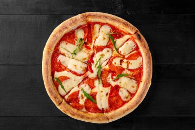 İnce dilimlenmiş salam sosisi, domates sosu, erimiş mozzarella peyniri ve siyah arka planda taze roka ile kaplanmış kahverengi hamurlu pizza. İtalyan-Amerikan mutfağı