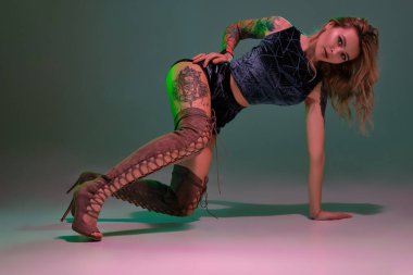 Dövmeli ve uzun kıvırcık saçlı çekici sarışın kadın kıvırcık dans ediyor ve pembe, gri arka planı ve kopyalama alanı olan beyaz bir arkaplanda kameraya bakıyor. Karanlıkta giyinmiş tatlı bir kız.