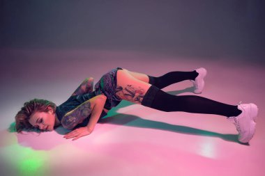 Young beautiful caucasian blonde girl with tattoo on her body, wearing velour blue booty shorts and black stockings dancing twerk lying down with raised booty, pink background