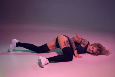 Young beautiful caucasian blonde girl with tattoo on her body, wearing velour blue booty shorts and black stockings dancing twerk lying down with raised booty, pink background