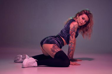 Young beautiful caucasian blonde girl with tattoo on her body, wearing velour blue booty shorts and black stockings dancing twerk on her knees, pink background