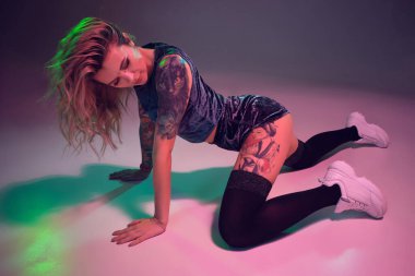 Young beautiful caucasian blonde girl with tattoo on her body, wearing velour blue booty shorts and black stockings dancing twerk on her knees, pink background
