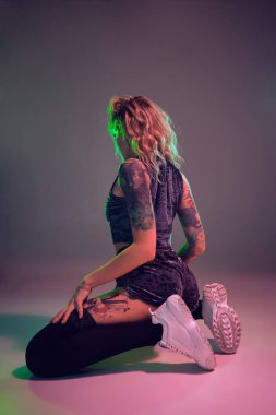 Young beautiful caucasian blonde girl with tattoo on her body, wearing velour blue booty shorts and black stockings dancing twerk lying down with raised booty, pink background