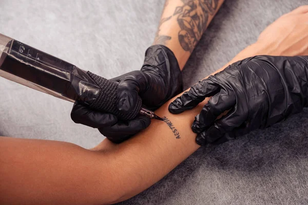 タトゥーの作成をレタリングするプロセス 黒いゴム製手袋のプロのタトゥーアーティストの手 巧みにマシンを使用して クライアントの前腕にアエステティックという単語をエッチング クロップショット — ストック写真