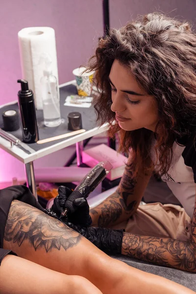 才能のある若い女性のタトゥーアーティストは クライアントの太ももにユニークな花のデザインをもたらすために機械を使用してサロンで少女のためのタトゥーを勤勉に作っている見ました — ストック写真