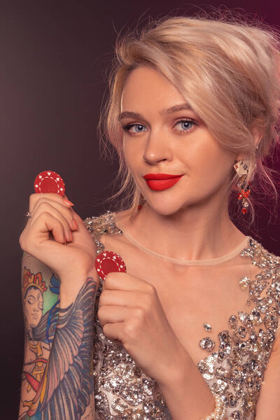 Крупный план очаровательной леди с светлыми волосами, татуированными руками и идеальным макияжем, одетой в серебряное блестящее платье, позирующей с красными фишками в руках и улыбающейся. Концепция покера на