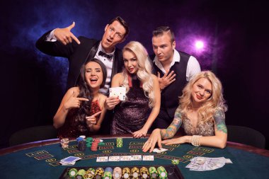 Bir grup komik genç arkadaş kumarhanede poker oynuyorlar. Gençler büyük bir galibiyet için bahis oynuyorlar. Gülümsüyorlar ve masada kırmızı ve mavi arka farlara karşı poz verirken heyecanlı görünüyorlar.