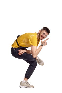Gözlüklü komik bir dansçının tam boy portresi, siyah tulum, sarı tişört ve stüdyoda aylak aylak dolaşan gri spor ayakkabılar. Beyaz arka planda dans eden elementleri izole eden bir adamın kapalı alanda çekilmiş fotoğrafı.